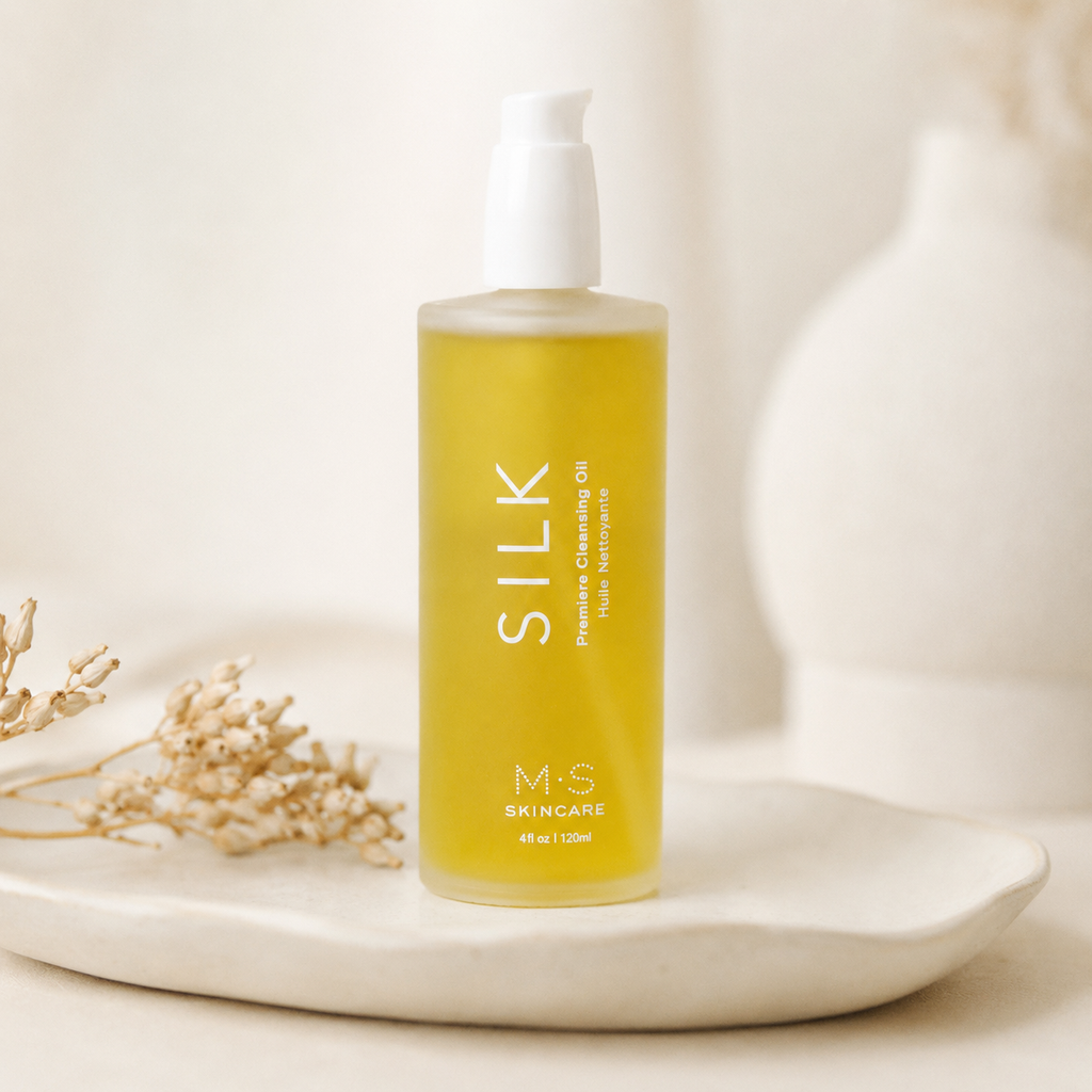 A bottle of M-S Skincare Silk Purifying Cleansing Oil with a white pump dispenser, filled with golden-yellow oil, displayed on a white ceramic tray with dried flowers against a soft, warm beige background.