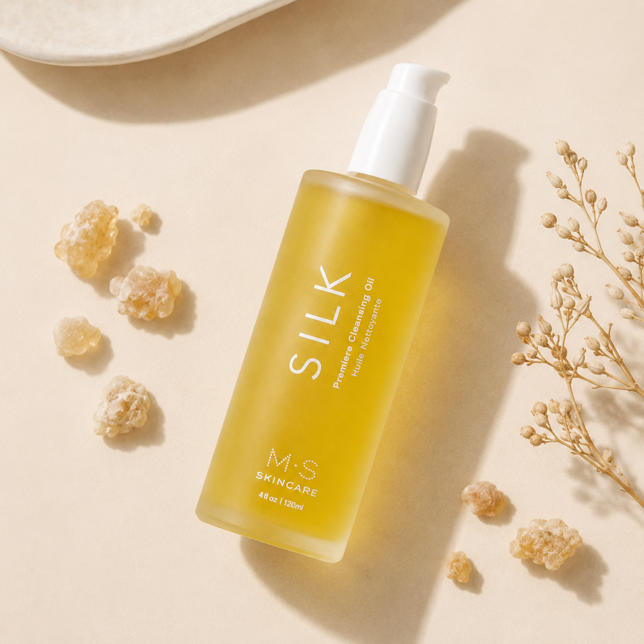 A bottle of M-S Skincare Silk Retinol Cleansing Oil with a white pump dispenser, filled with golden-yellow oil, styled on a warm beige background with dried flowers and crystals.