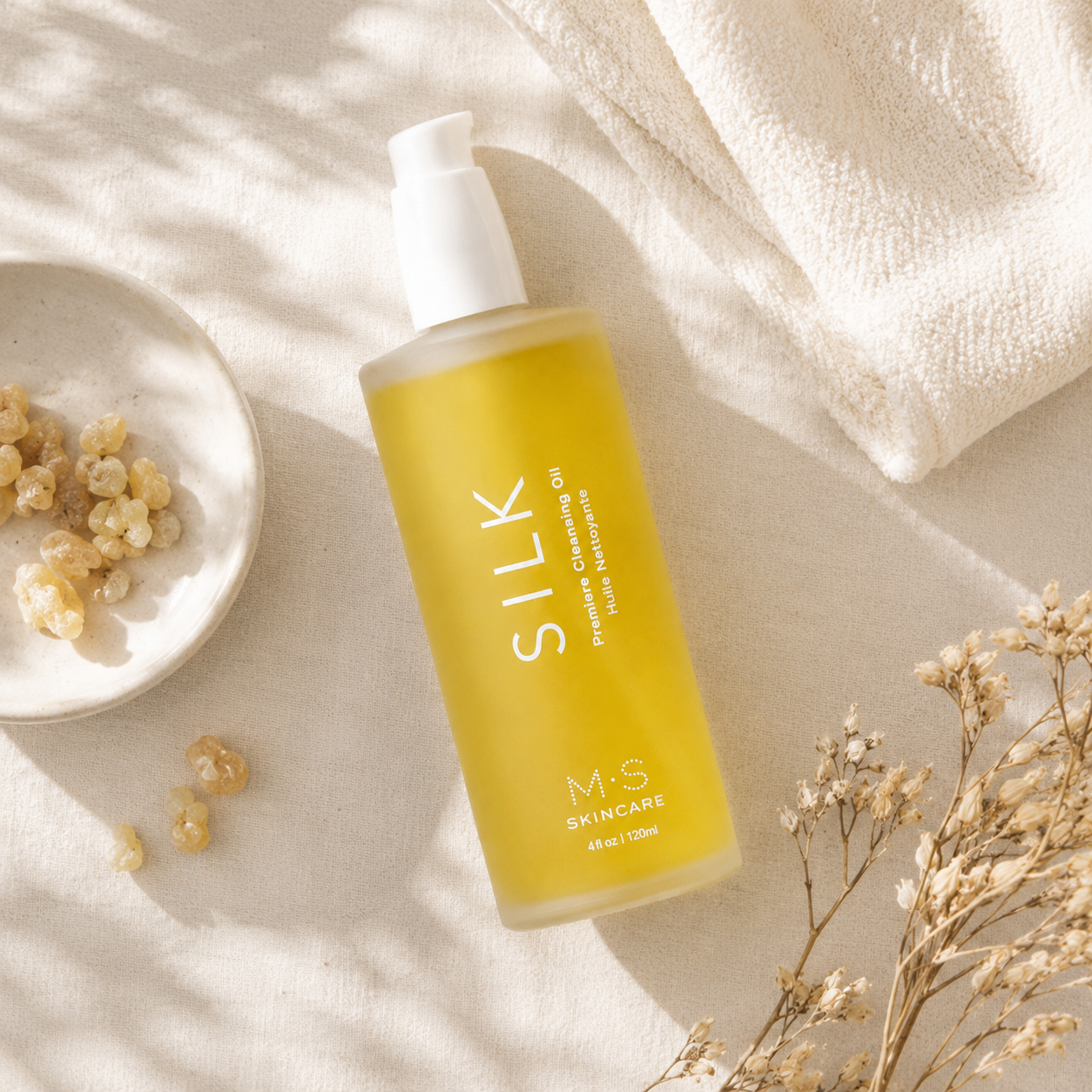 M.S Skincare Silk Phyto-Rejuvenating Cleansing Oil in a frosted bottle with a white pump, styled on a beige linen surface with dried flowers, a small dish of botanicals, and a folded cream towel.