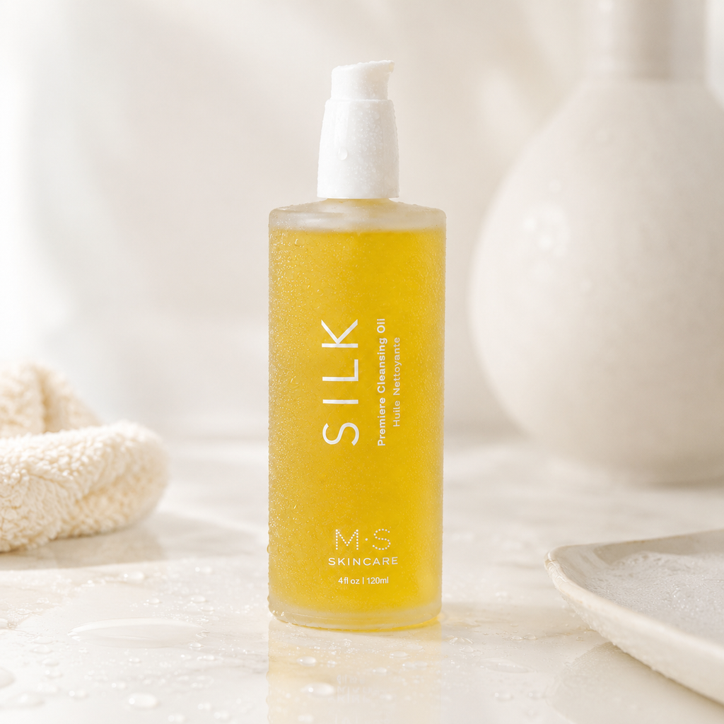 A bottle of M-S Skincare Silk Premium Cleansing Oil with a white pump dispenser, filled with golden yellow oil, set on a wet marble surface with water droplets, surrounded by soft, warm neutral props.