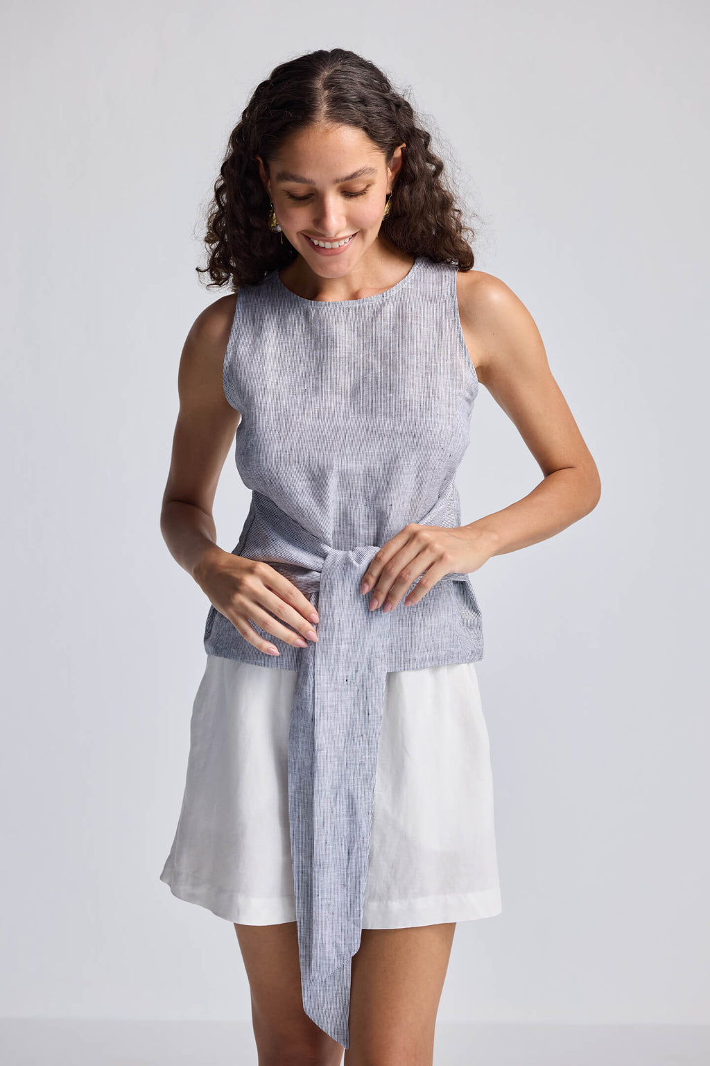 Woman wearing a sleeveless gray top with a tie waist and white shorts on a light gray background
