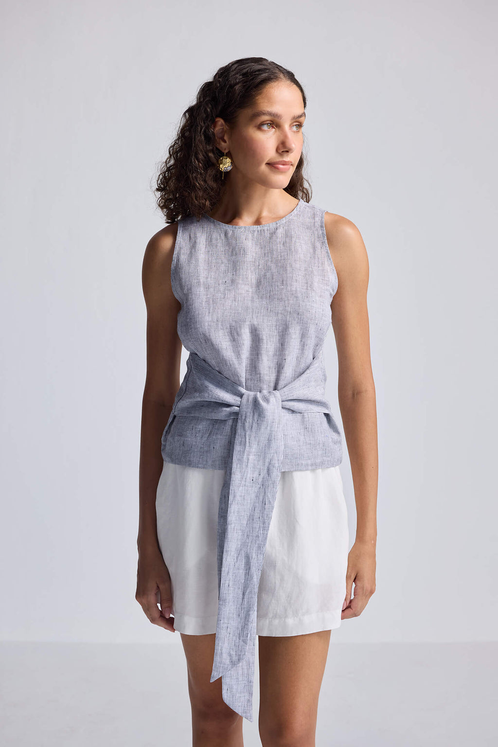 Woman wearing a sleeveless top with a tie front and white shorts on a plain background