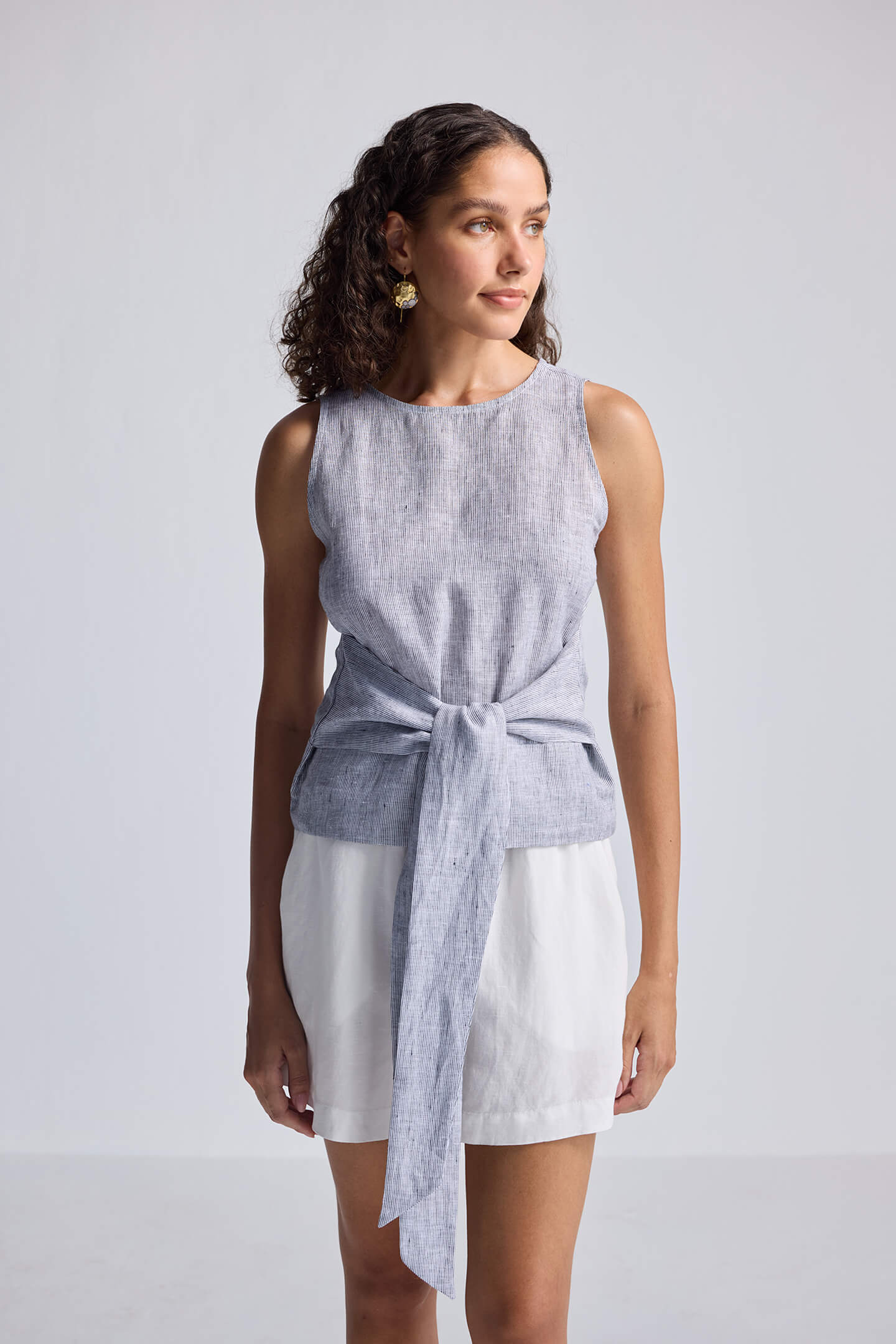 Woman wearing a sleeveless top with a tie front and white shorts on a plain background