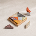 A geometric wooden puzzle set with triangular stone tiles in earthy colors, arranged on a small wooden tray with a few loose pieces on a light beige textured surface.
