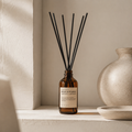An amber glass reed diffuser bottle with a cream label and several black reed sticks, placed on a light stone ledge beside a textured ceramic vase.