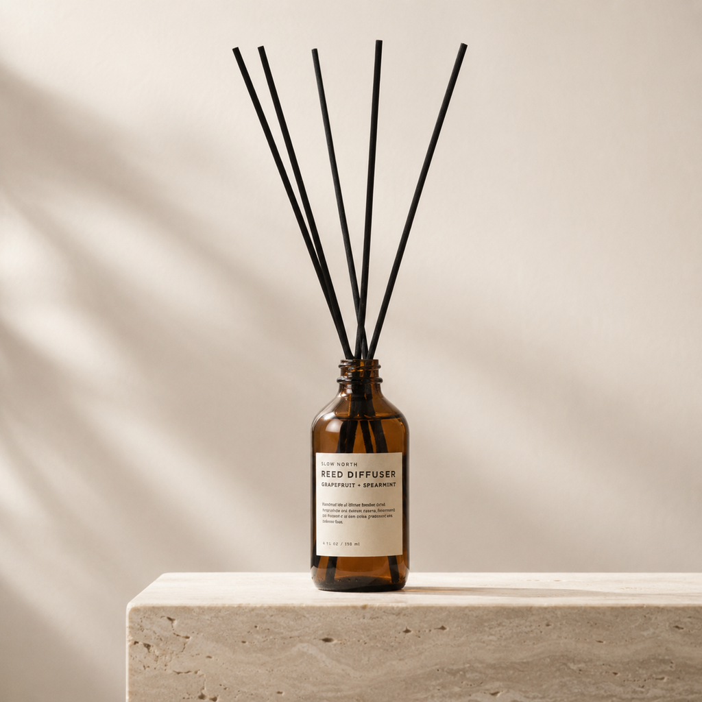 Amber glass reed diffuser bottle with a cream label and six black reed sticks, displayed on a light stone pedestal against a warm beige wall with soft leaf shadows.