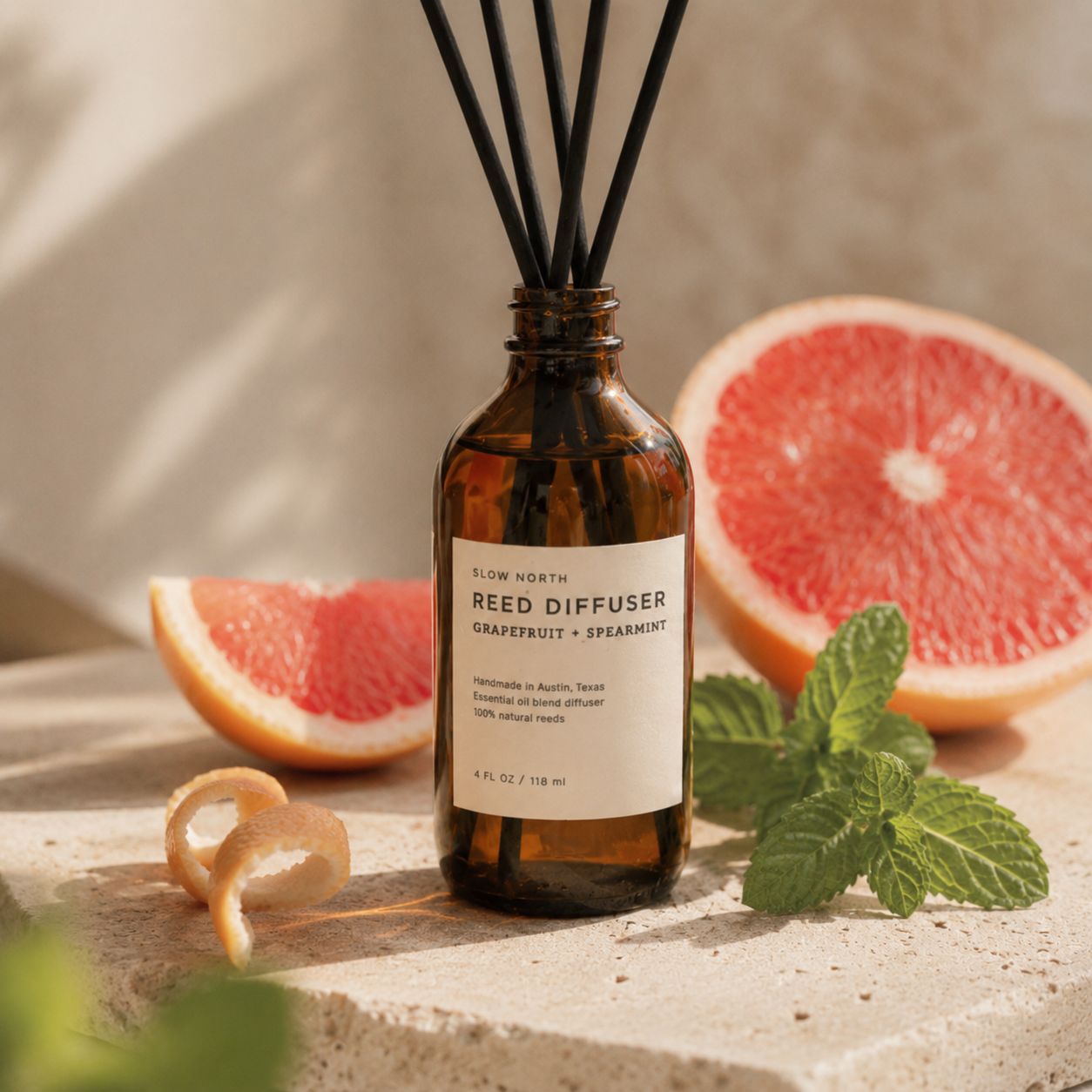 An amber glass reed diffuser bottle with a white label reading 'SLOW NORTH REED DIFFUSER GRAPEFRUIT + SPEARMINT,' styled with grapefruit slices, mint leaves, and a citrus peel on a stone surface.