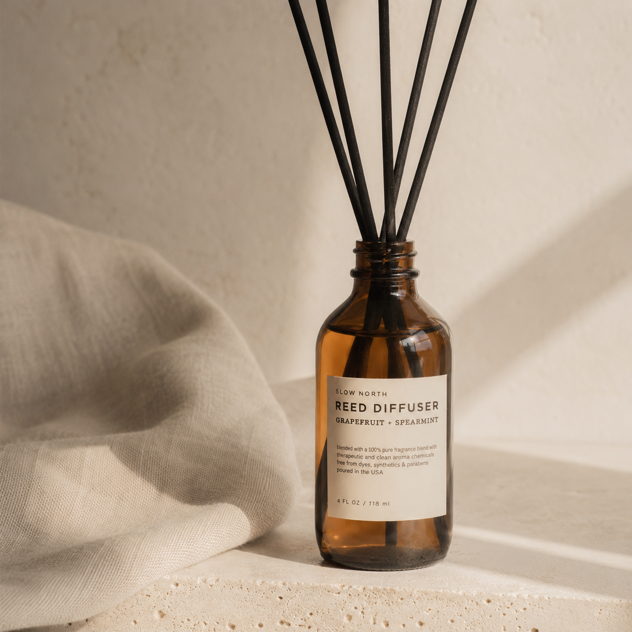 Amber glass reed diffuser bottle with black reed sticks and a cream label reading 'Reed Diffuser - Grapefruit + Spaghetti' by New North, styled on a textured surface with a draped linen cloth.