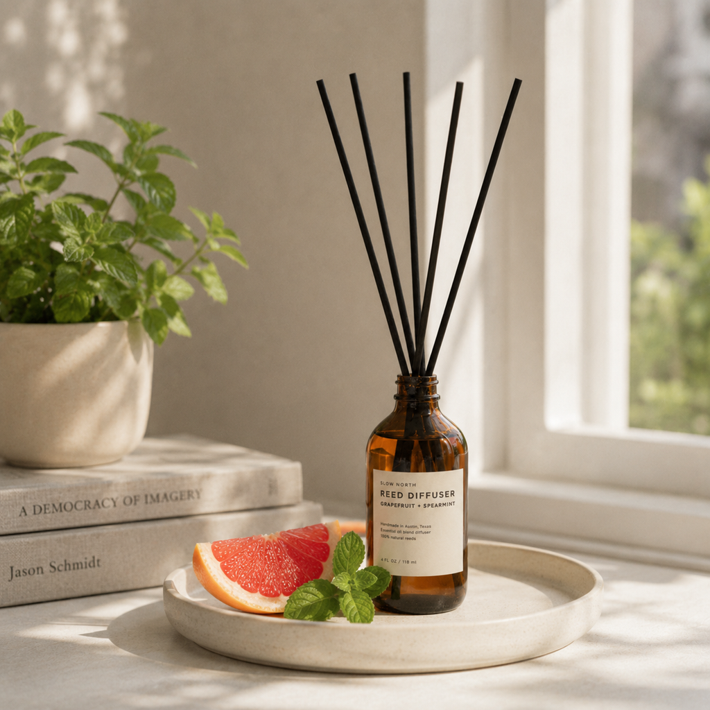 An amber glass reed diffuser bottle with black reeds stands on a round tray beside grapefruit slices and mint leaves in a bright indoor setting near a window.