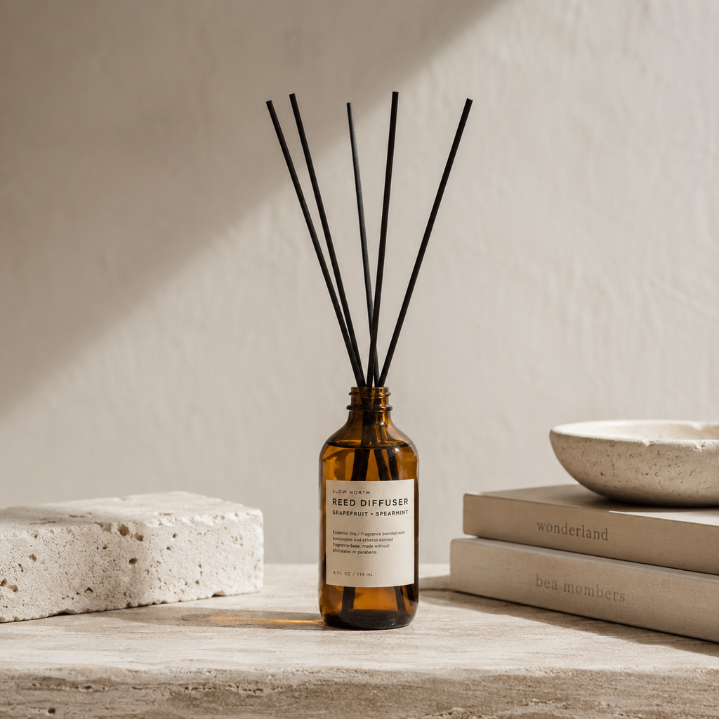 Amber glass reed diffuser bottle with a white label and six black reeds, styled on a stone surface with books and a ceramic bowl against a textured beige wall.