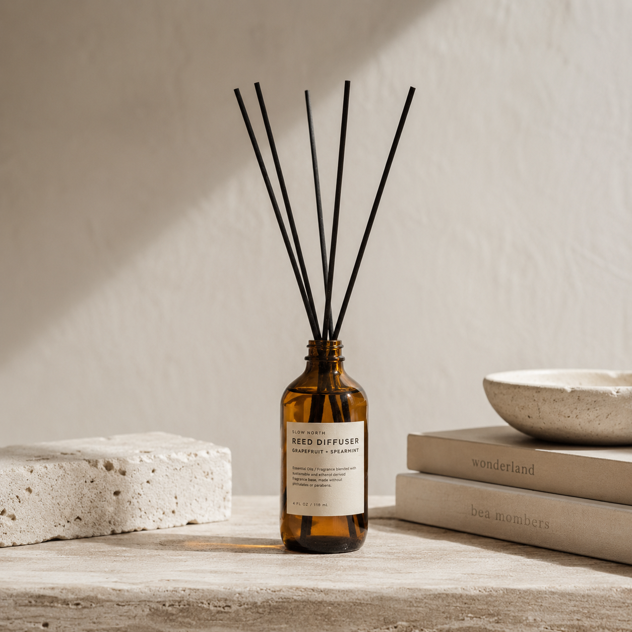 Amber glass reed diffuser bottle with a white label and six black reeds, styled on a stone surface with books and a ceramic bowl against a textured beige wall.