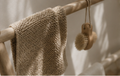 A beige knitted washcloth hangs from a wooden rod next to a wooden scrubber with natural bristles, set against a softly lit neutral background.