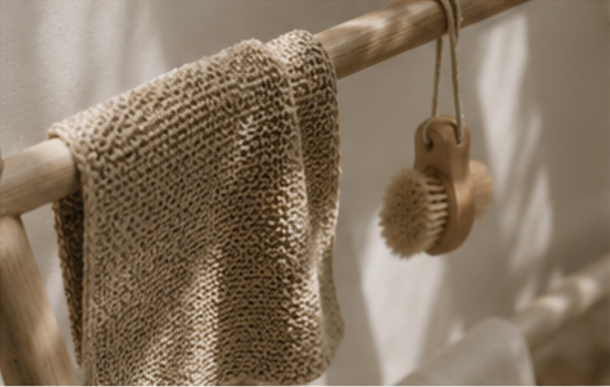 A beige knitted washcloth hangs from a wooden rod next to a wooden scrubber with natural bristles, set against a softly lit neutral background.