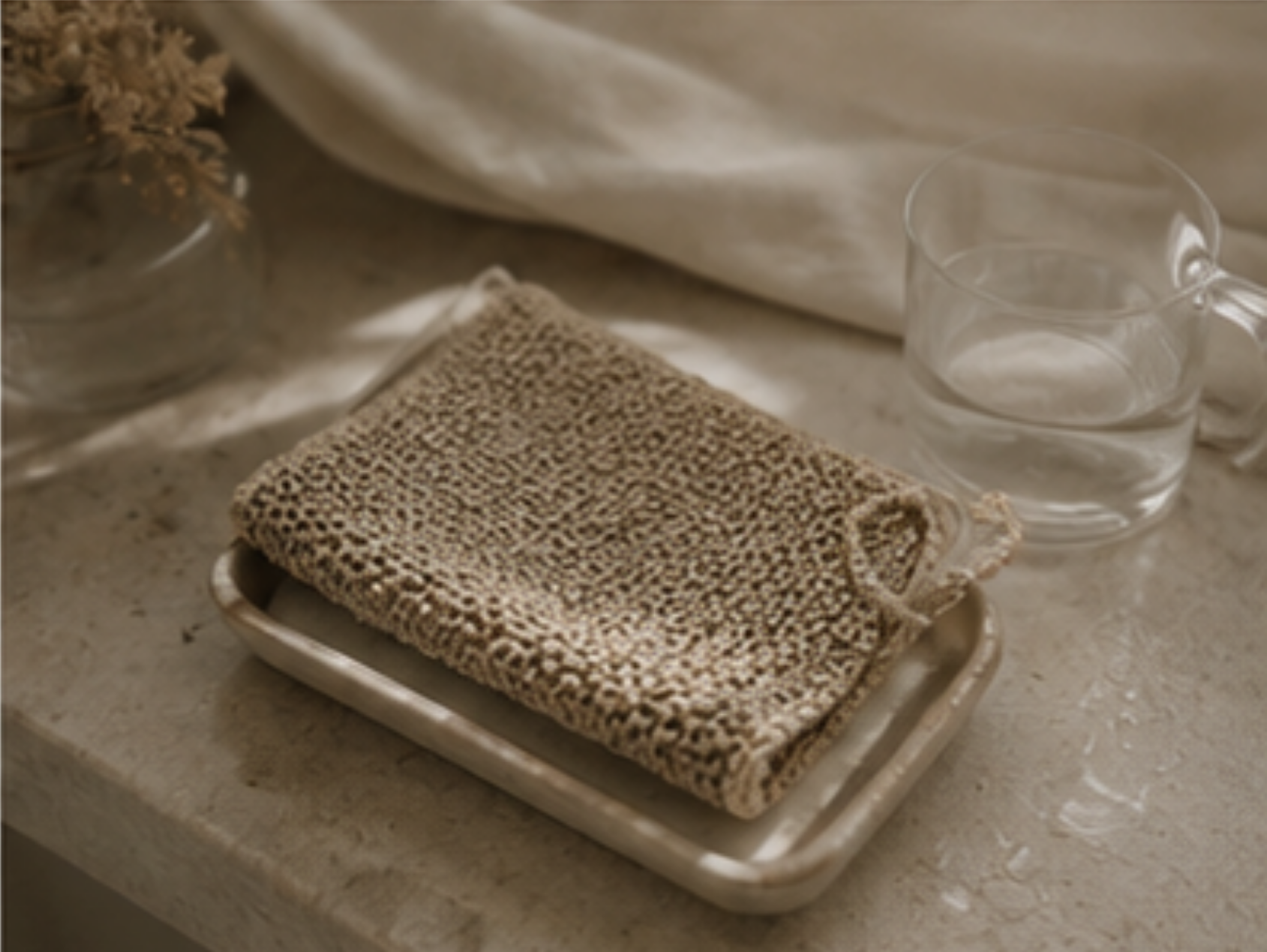 A beige woven scrubber or cleaning cloth with a loop handle, resting on a small metal tray next to a clear glass cup on a stone surface.