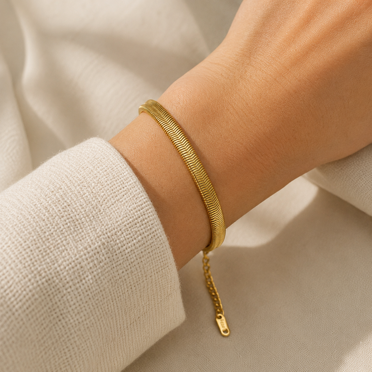 A close-up of a gold bangle bracelet with a textured, snake-like pattern and a small hanging chain clasp, worn on a person's wrist against a soft, off-white background.