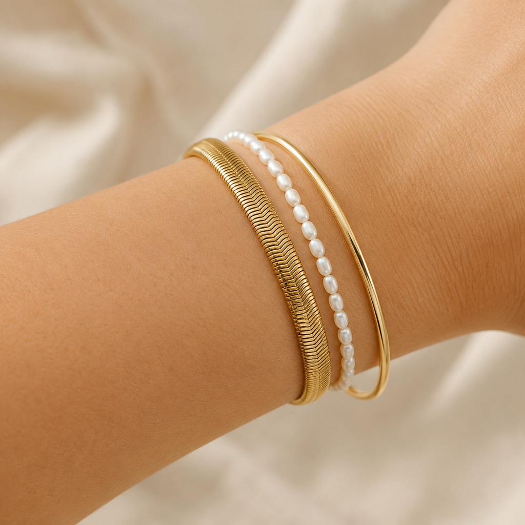 Three delicate bangles worn on a wrist: a textured gold snake chain bracelet, a smooth gold bangle, and a strand of white pearl beads.