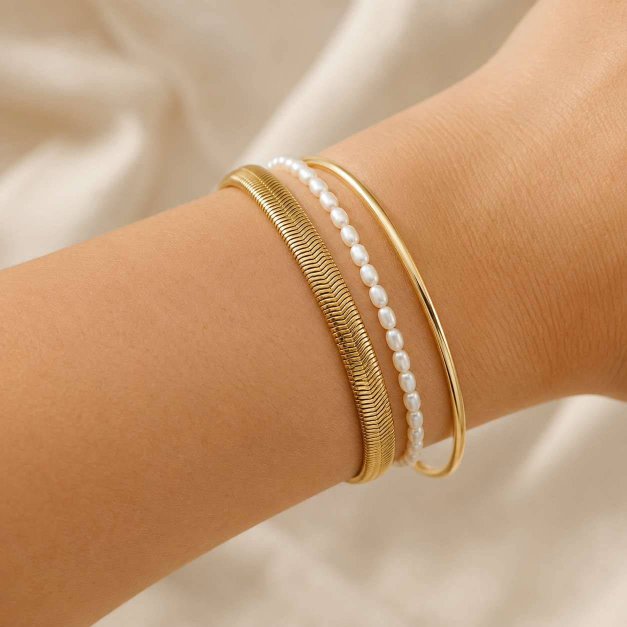 Three delicate bangles worn on a wrist: a textured gold snake chain bracelet, a smooth gold bangle, and a strand of white pearl beads.