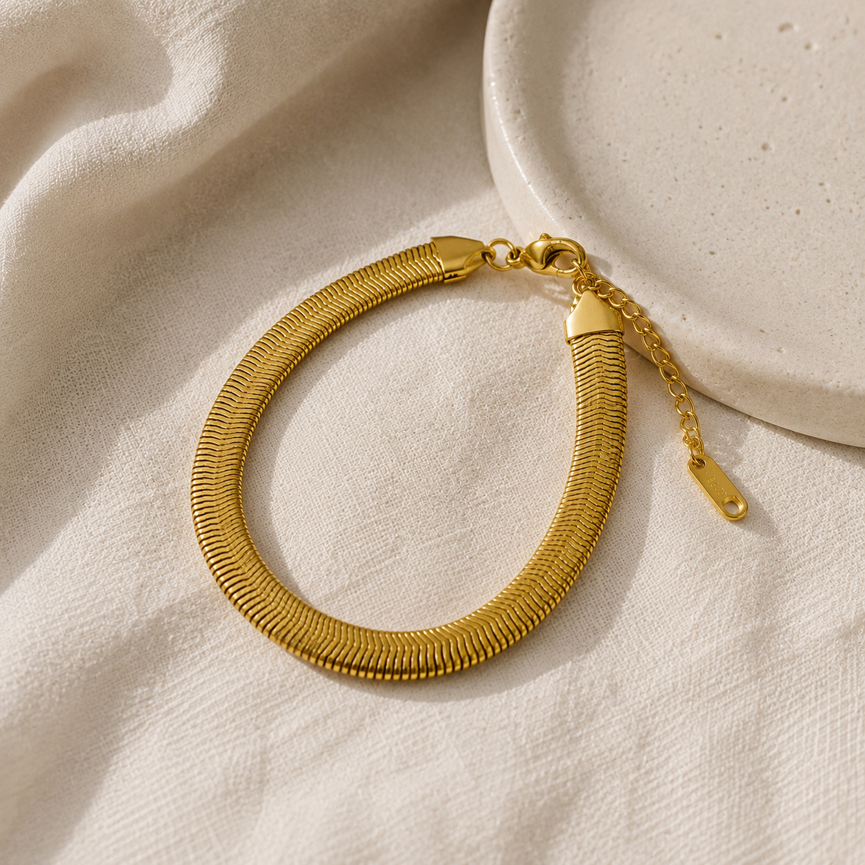 A gold-toned snake chain bracelet with a lobster clasp and extension chain, styled on draped cream fabric next to a textured ceramic dish.
