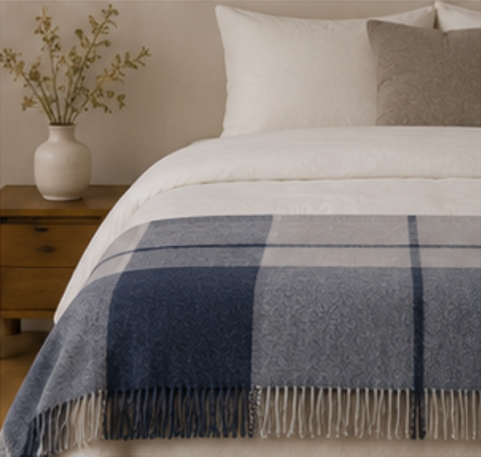 A blue and gray plaid throw blanket with fringe is draped over a bed with white bedding, next to a wooden nightstand with a vase of dried branches.