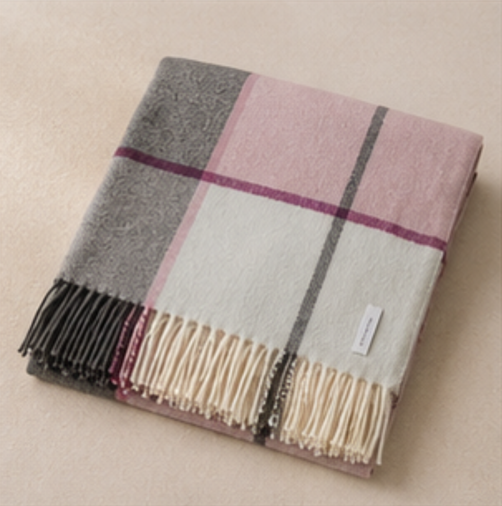 A folded plaid throw blanket with cream, dusty pink, gray, and dark purple stripes, featuring cream fringe and a small white brand tag, set against a neutral beige background.