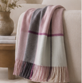 A soft woven throw blanket draped over a cream sofa, featuring a plaid pattern in dusty pink, charcoal gray, and off-white with fringe at the ends. A small vase and books sit on a wooden side table in the softly lit background.