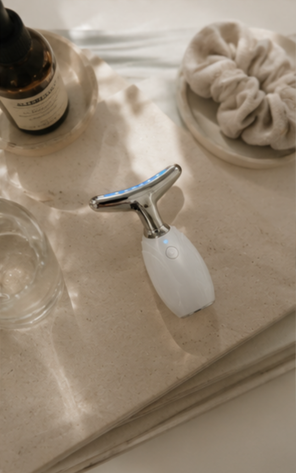A handheld beauty device with a white handle and silver metallic head, resting on a beige stone surface with styled bathroom props including a bottle, glass, and scrunchie.