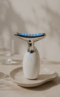 A handheld beauty device with a white body and a curved silver metallic head featuring blue LED lights, resting on a textured beige ceramic dish against a warm, softly lit background.