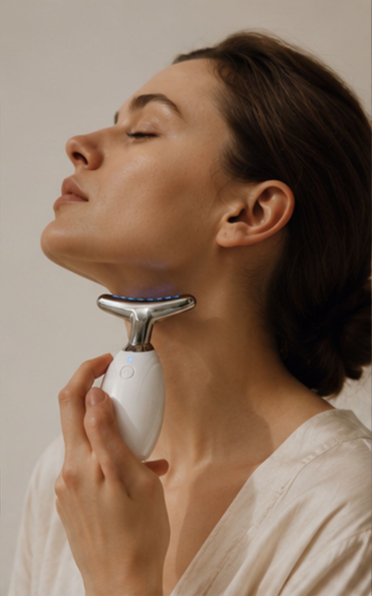 A woman with her eyes closed uses an electric facial skin-tightening device on her neck, wearing a white robe against a neutral beige background.