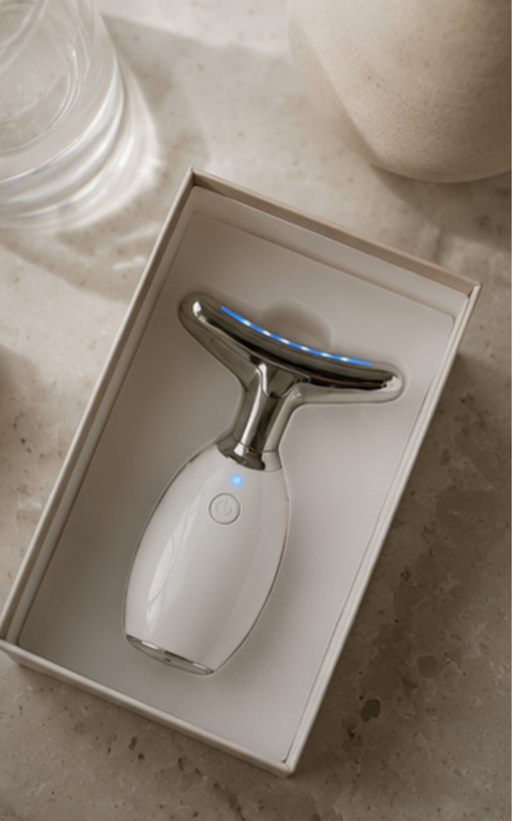 A white handheld beauty device with a metallic silver T-shaped head and blue LED light strip, presented inside an open white box on a light stone surface.
