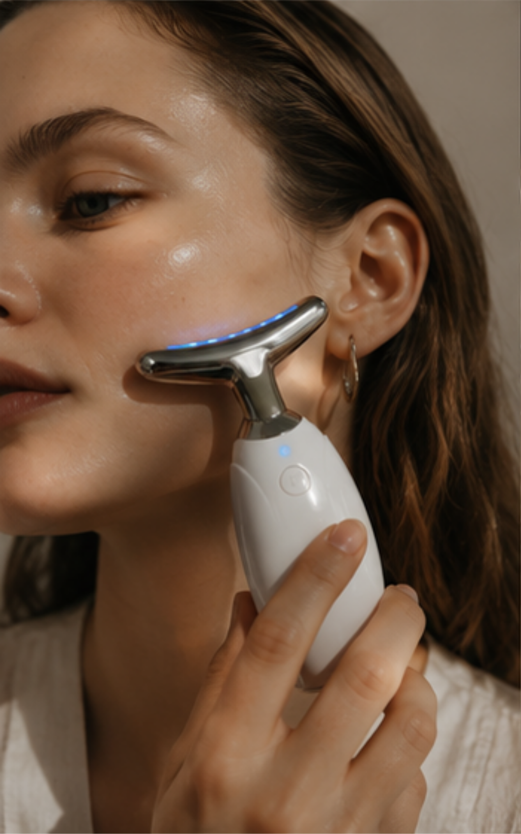 A close-up of a woman using a white and silver handheld facial beauty device on her cheek, with warm lighting and a soft neutral background.