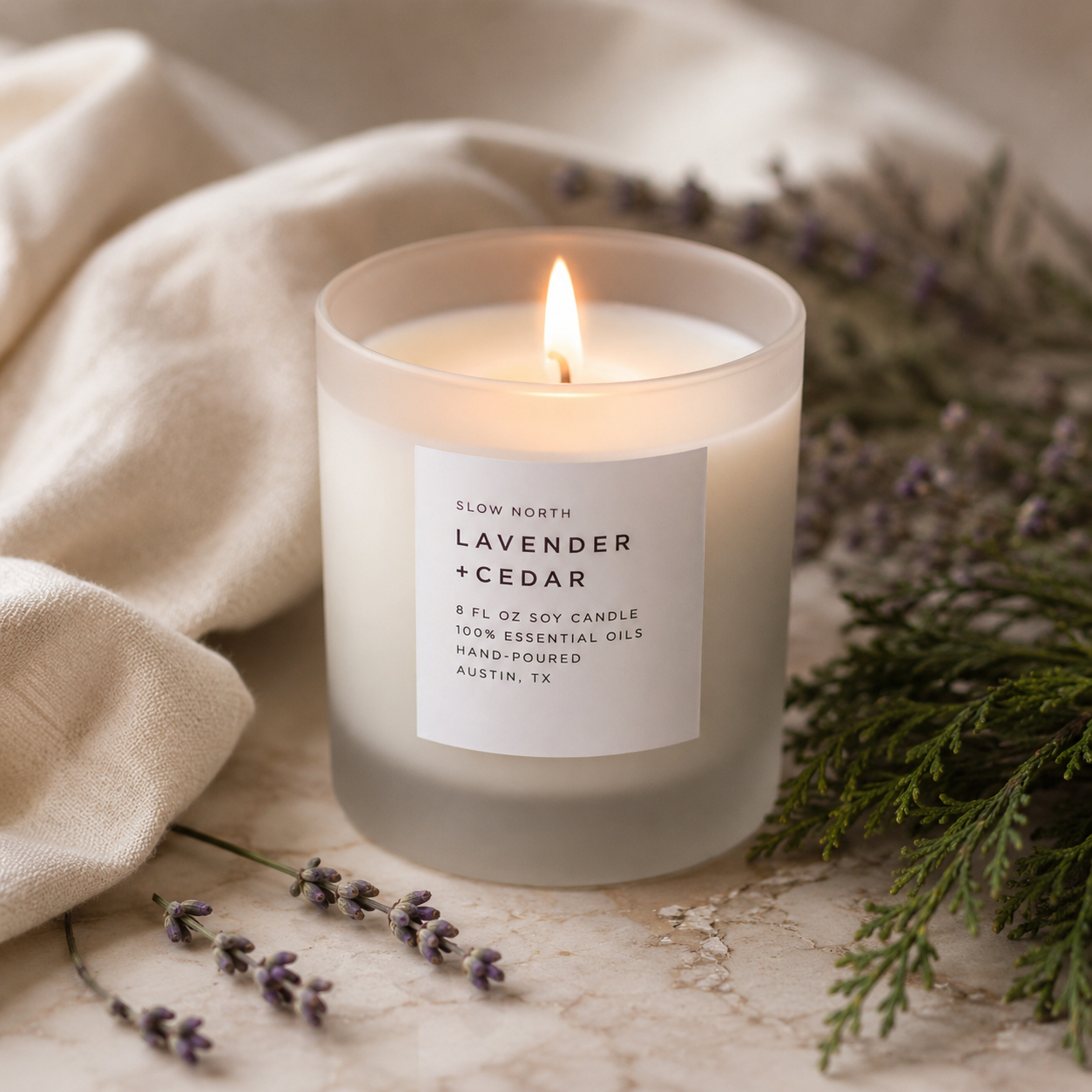 A lit soy candle in a frosted glass jar with a white label reading 'SLOW NORTH LAVENDER + CEDAR,' styled on a light stone surface with cream fabric, lavender sprigs, and greenery.