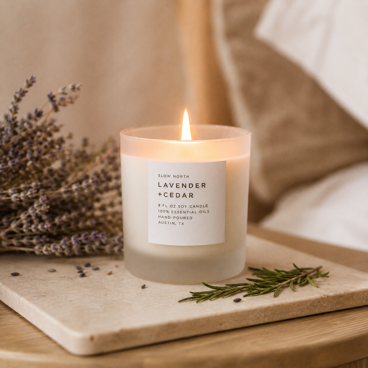 A lit soy candle in a frosted glass jar labeled 'Slow North Lavender + Cedar,' placed on a wooden surface with dried lavender and rosemary sprigs in a warm, softly styled setting.