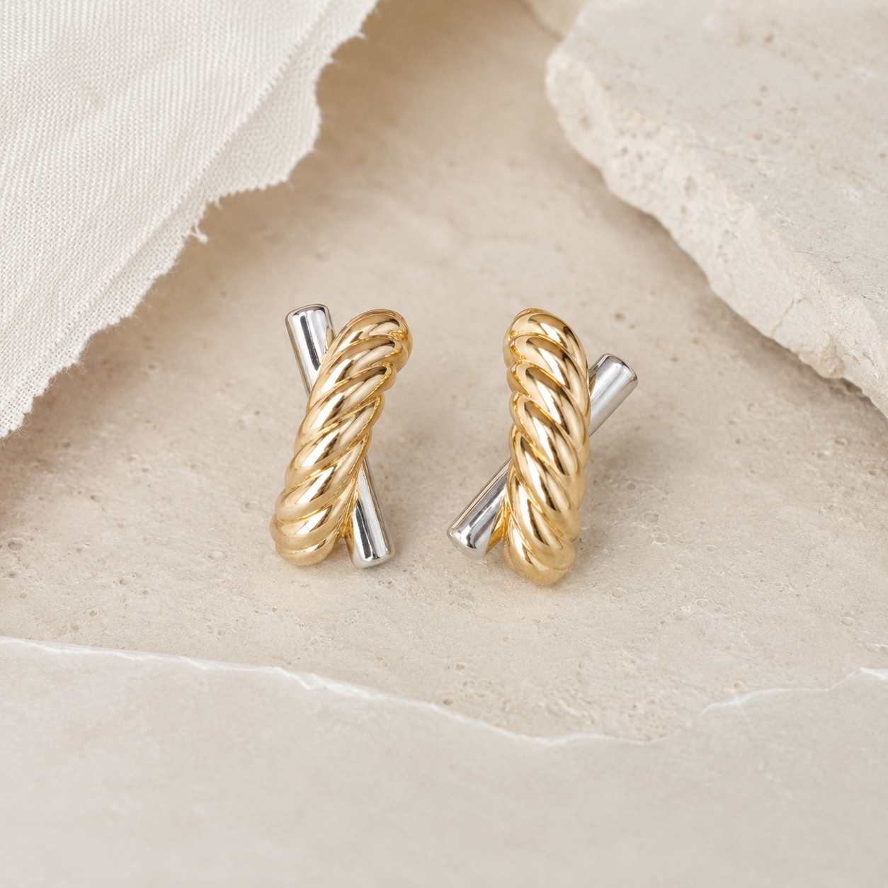 A pair of gold and silver twisted cable hoop earrings resting on a textured beige stone surface with a piece of white fabric in the background.