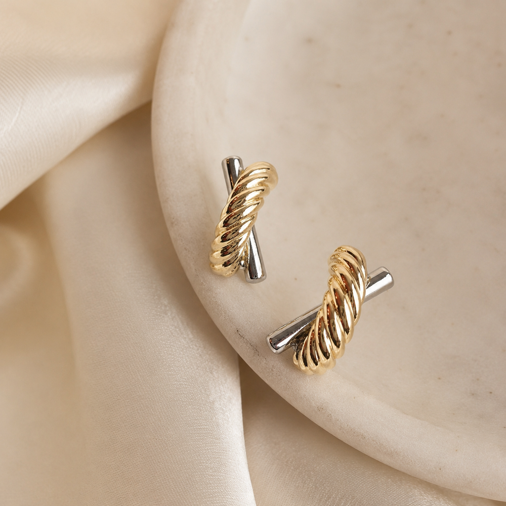 A pair of gold and silver twisted cable-style earrings resting on a cream-colored stone plate with draped satin fabric in the background.