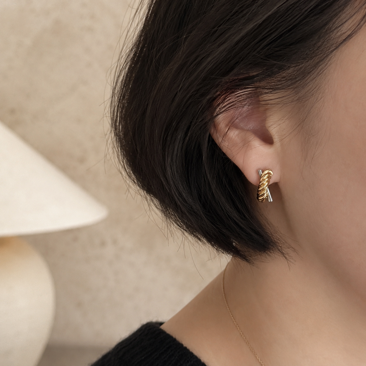 A close-up of a person's ear wearing a small twisted rope-style stud earring in yellow gold with a tiny silver accent, against a soft beige background.
