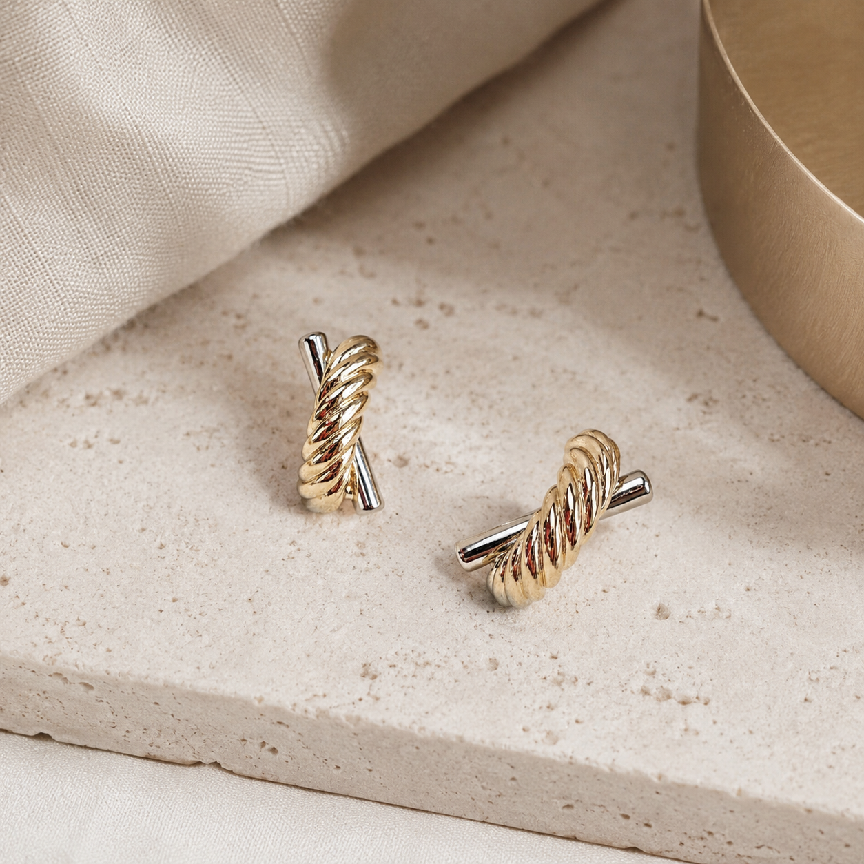 A pair of gold and silver twisted bar earrings displayed on a textured beige stone surface with a neutral fabric backdrop.