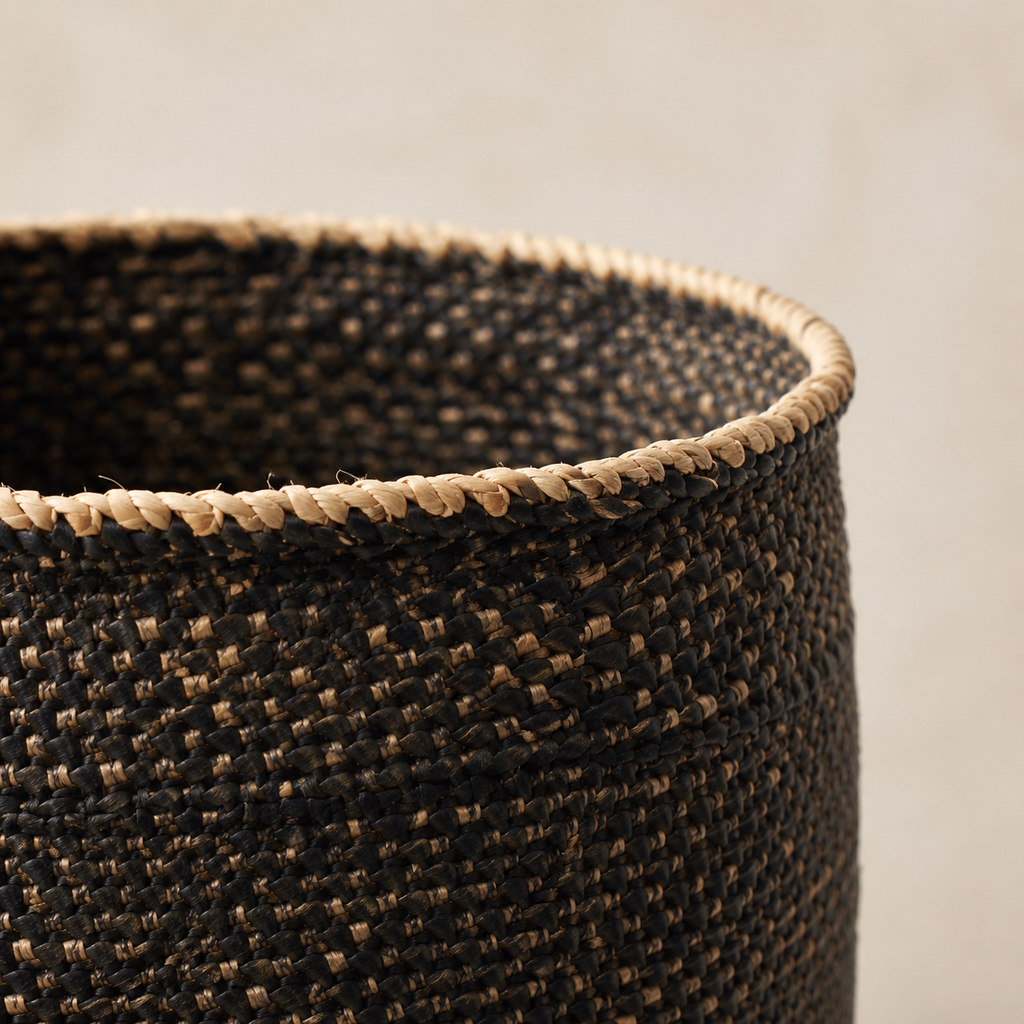 Close-up of a woven basket with a dark brown and black body and a natural beige rope trim along the rim, set against a neutral light gray background.