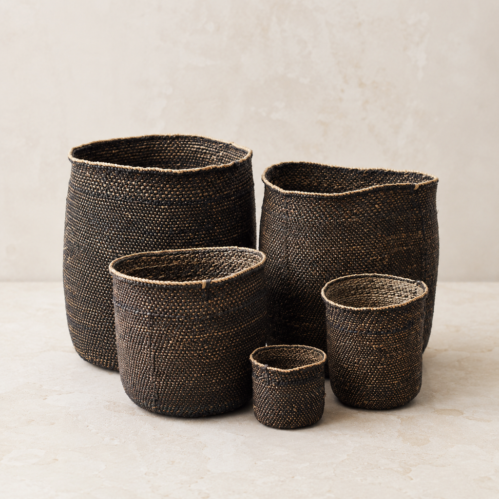 Four dark brown woven basket planters in different sizes arranged on a light beige surface against a textured beige wall.