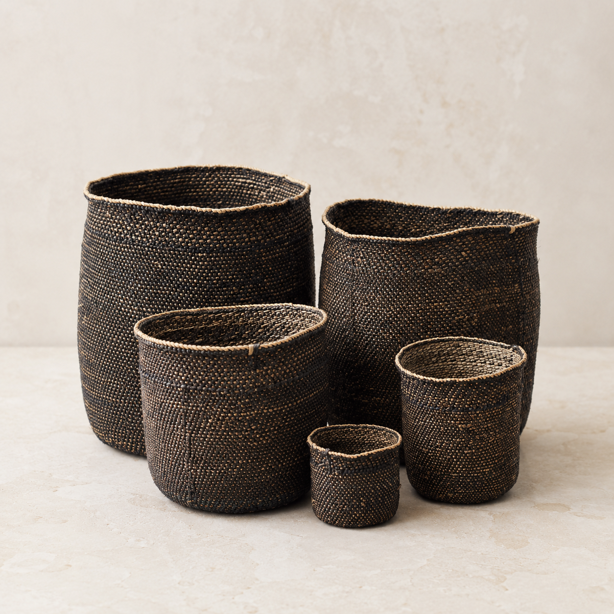 Four dark brown woven basket planters in different sizes arranged on a light beige surface against a textured beige wall.
