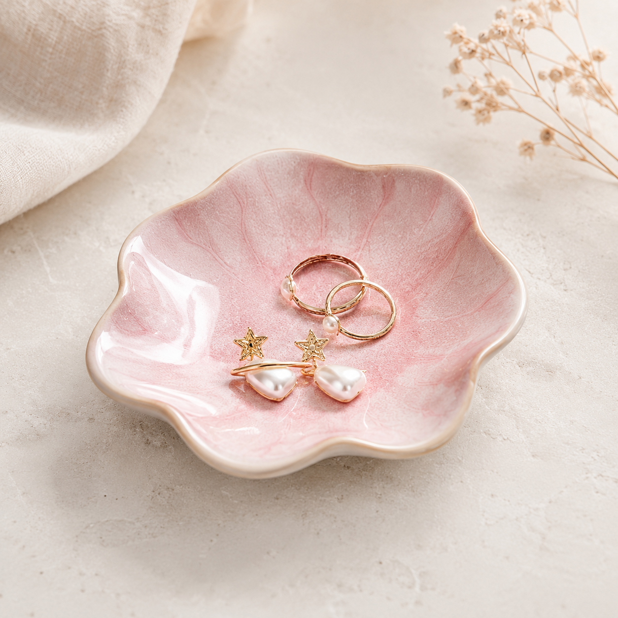 A pink marbled ceramic dish with a scalloped edge holds a pair of gold rings and pearl drop earrings with star charms, styled on a light textured surface with soft fabric and dried flowers.