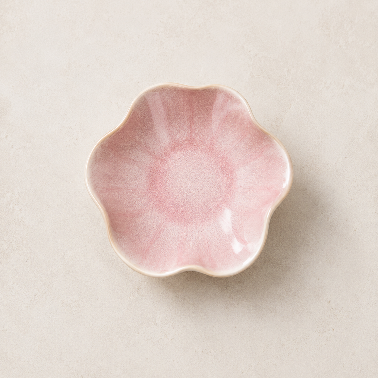 A small pink ceramic dish with a scalloped, flower-like rim, photographed from above on a light beige textured background.