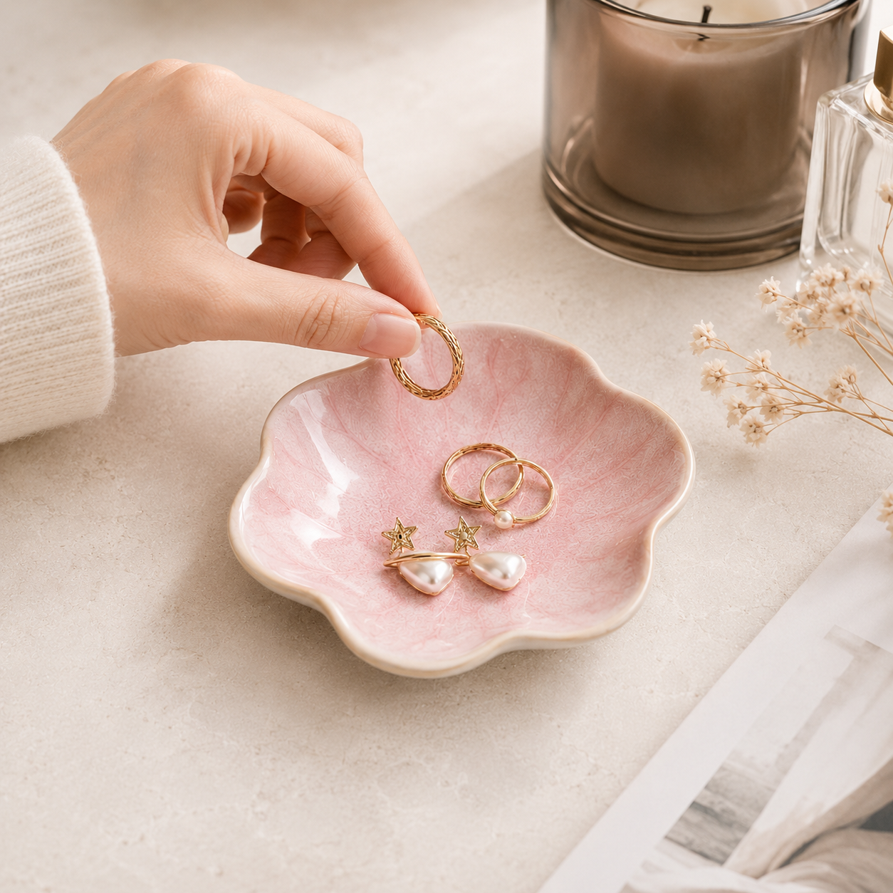 A hand wearing a white ribbed sweater places a gold ring into a small pink ceramic dish with a scalloped edge. The dish contains two gold rings and a pearl drop earring.