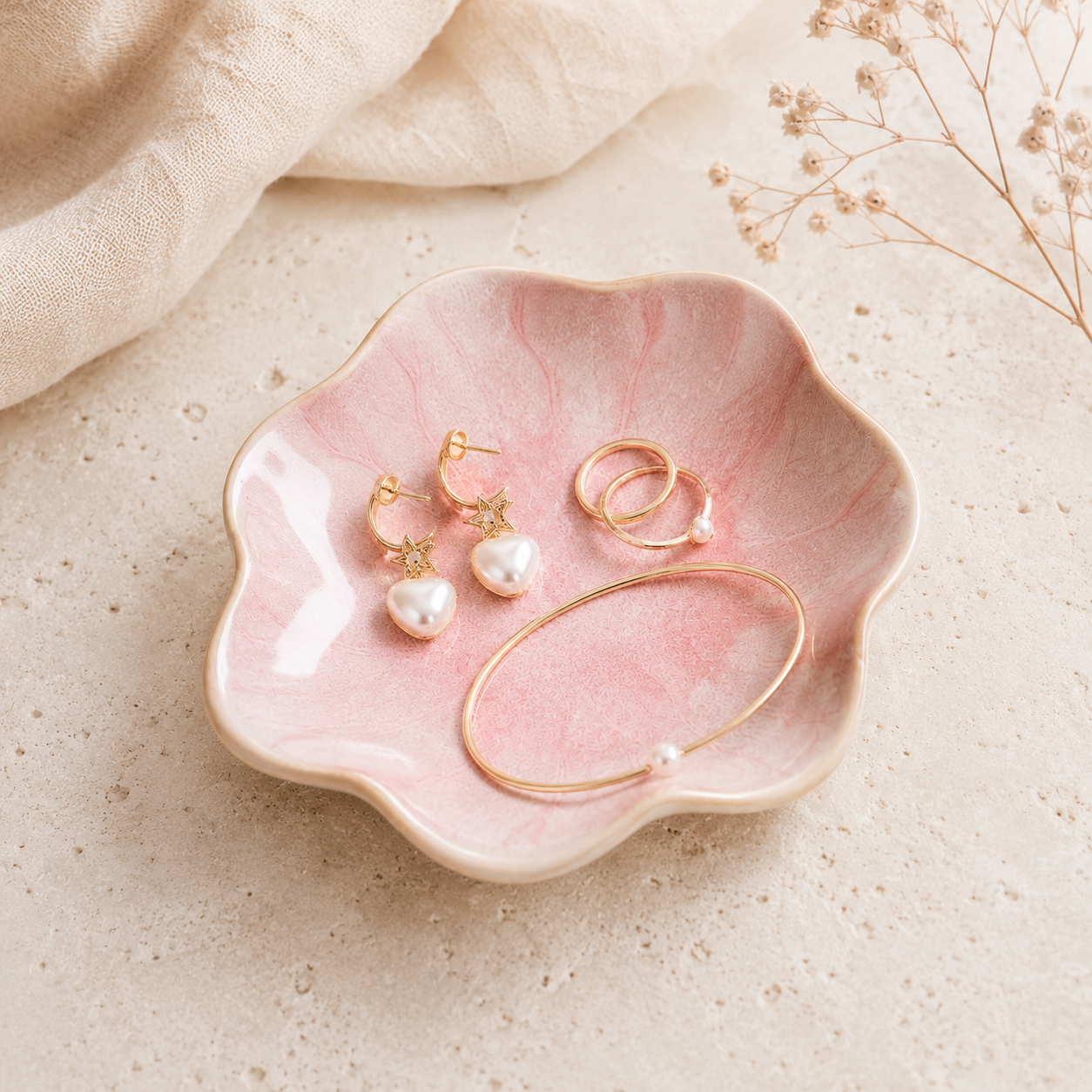 A pink marbled ceramic flower-shaped dish holding a pair of gold hoop earrings, a gold huggie ring, and a gold bracelet with a pearl bead, styled on a textured beige surface with soft fabric and dried flowers.