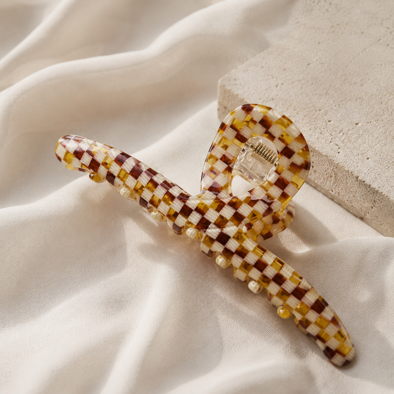 A checkered claw hair clip in amber, cream, and brown tones, decorated with small pearl-like beads, resting on draped cream fabric next to a textured stone block.