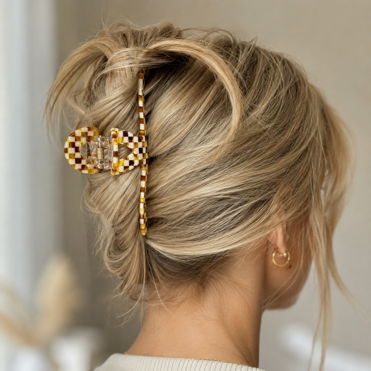 A blonde woman shown from the back wearing a large checkered claw hair clip in brown, yellow, and white, holding her hair in a messy bun.