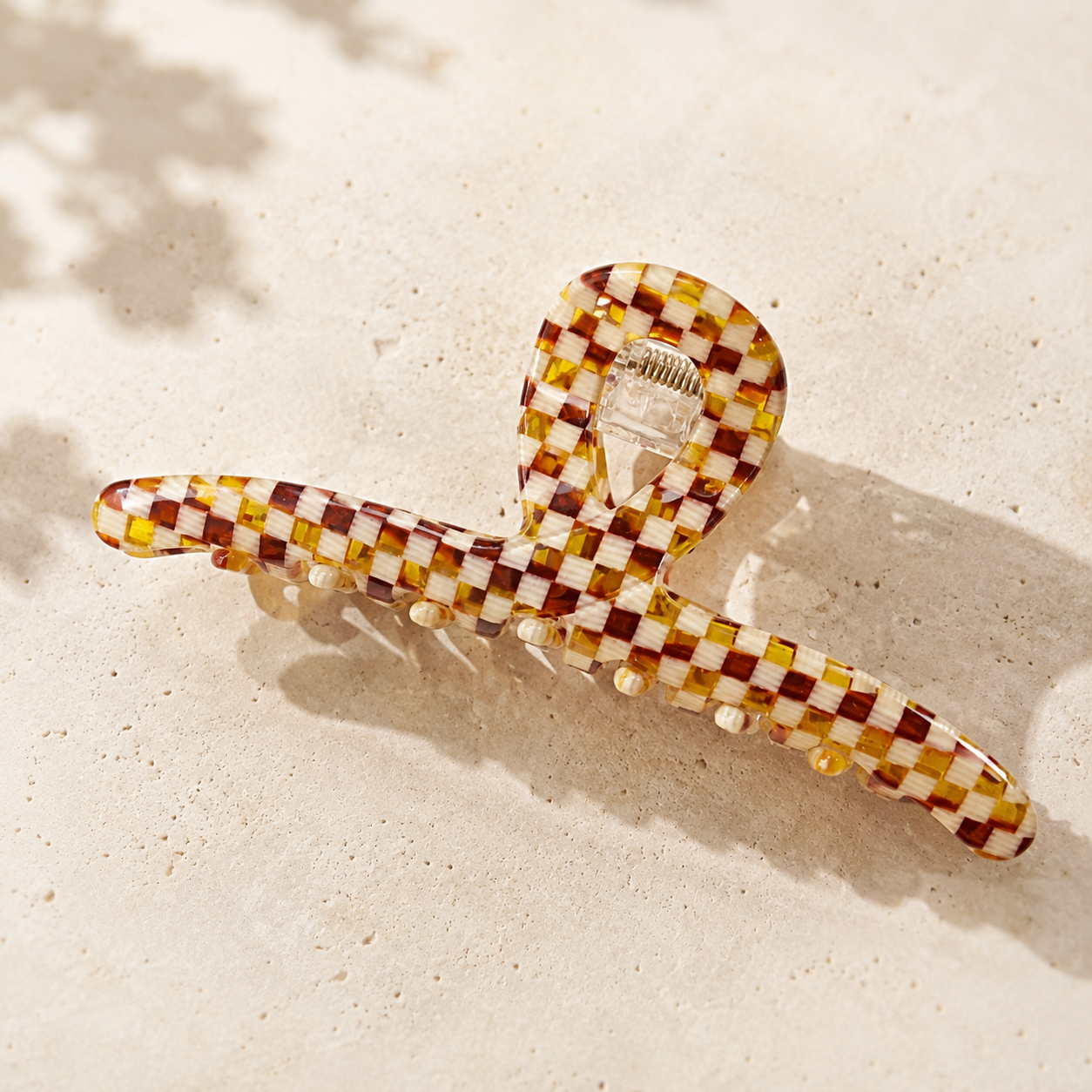 A translucent checkered hair claw clip with a brown, yellow, and white pattern, resting on a textured beige surface with soft shadows.