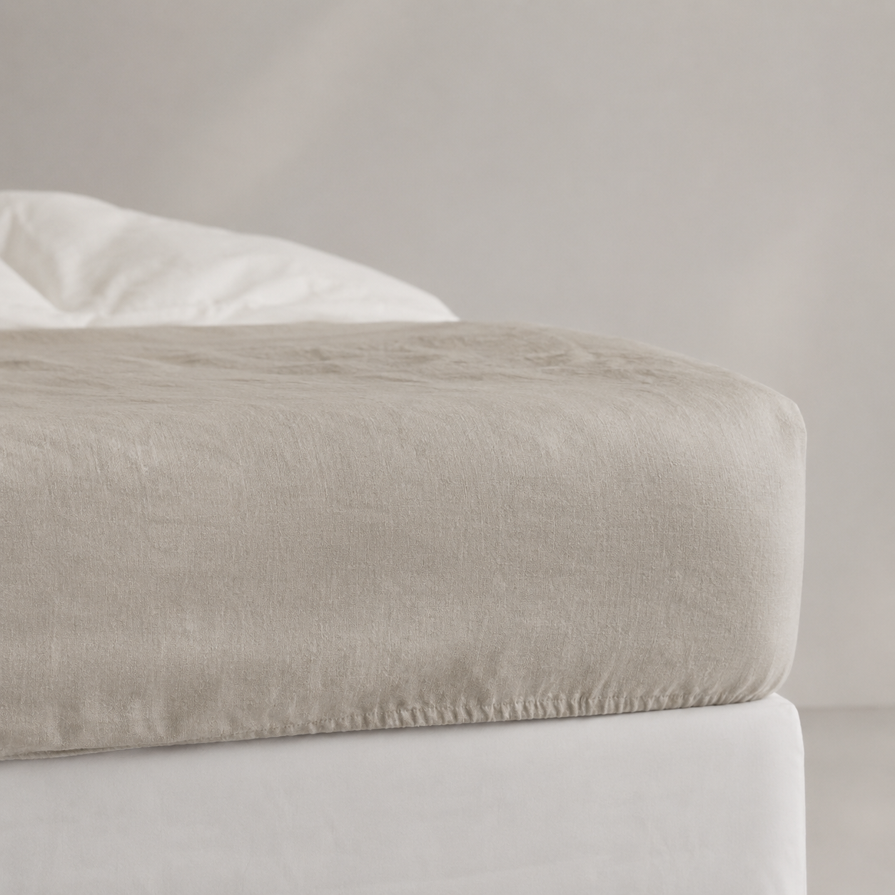 A close-up of a fitted sheet in a light taupe-beige color draped over the corner of a bed frame, with white bedding and pillows visible in the background.