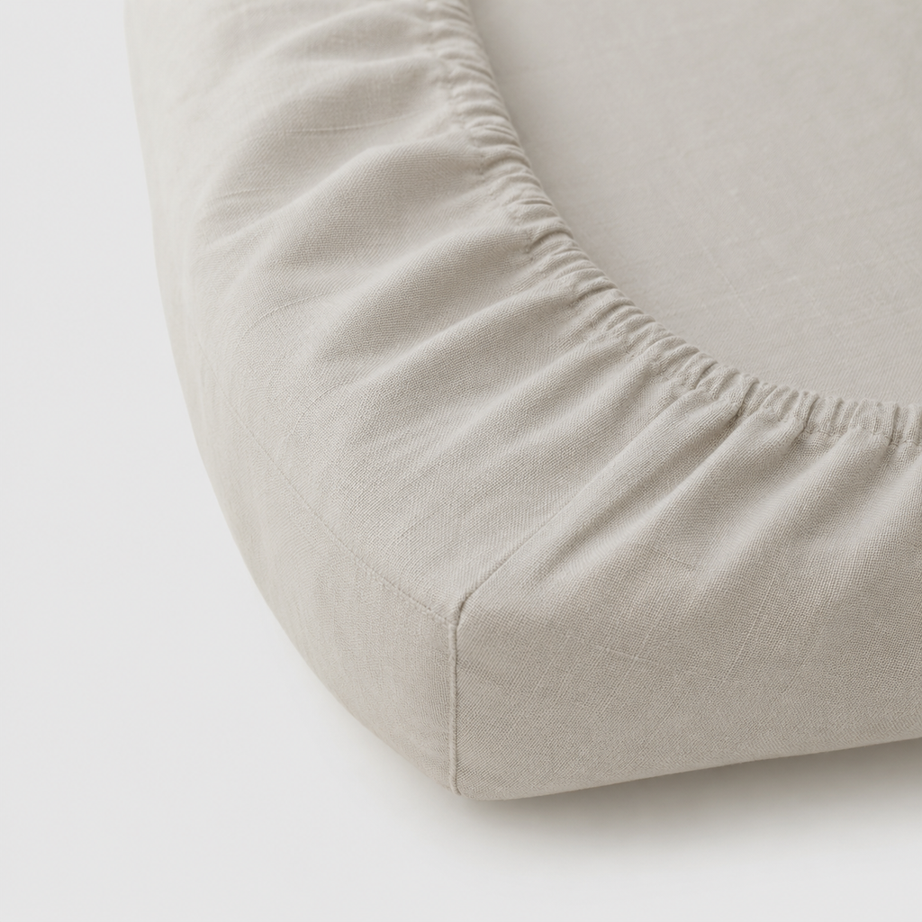 Close-up of a light beige fitted sheet with gathered elastic edges on a mattress corner, set against a plain white background.