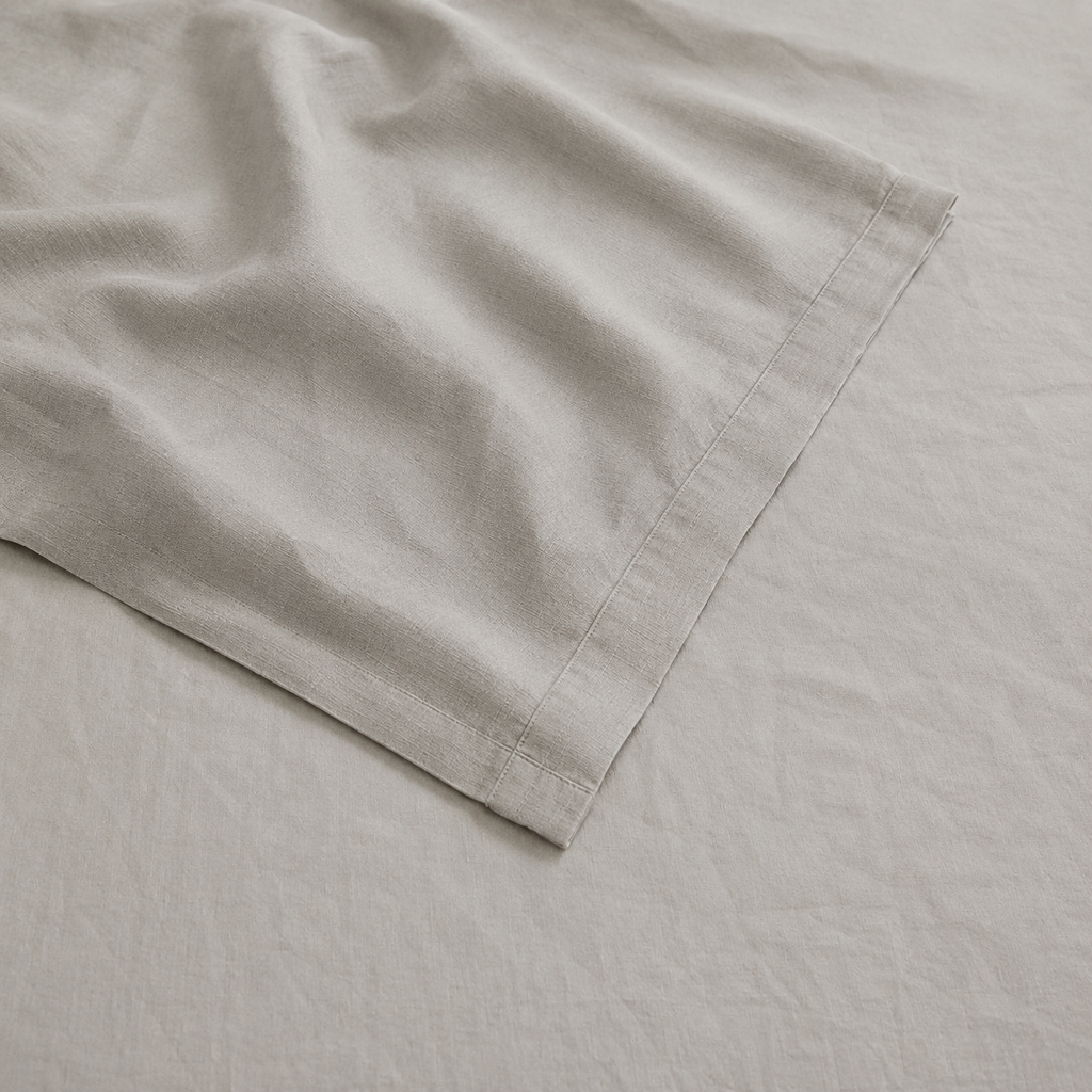 A close-up of a light beige fabric product, likely a linen sheet or pillowcase, draped diagonally across a matching textured fabric background.