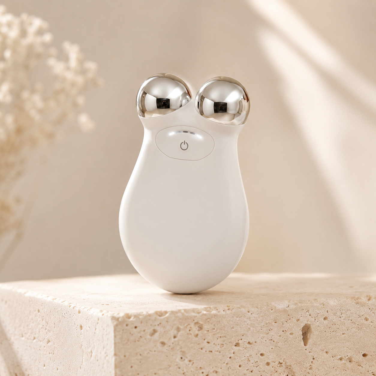 A white handheld beauty device with two shiny silver metal globes on top, shown standing on a light stone block against a soft beige background with dried flowers and sunlight.